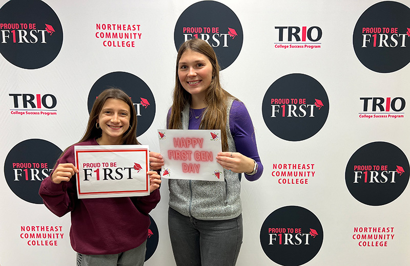2 students holding signs that say proud to be first and happy first gen day in front of trio backdrop