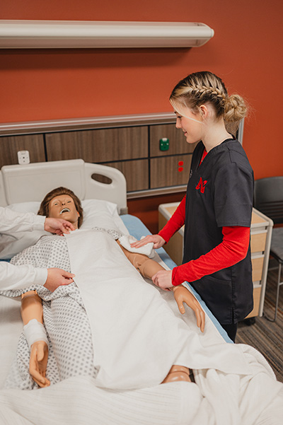 Students practicing patient care on a medical training manikin in a hospital bed.