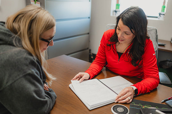 Two people sit at a desk reviewing a spiral‑bound document