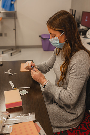 student wearing mask and gloves holding vial with needle and simulated skin injection pad on table