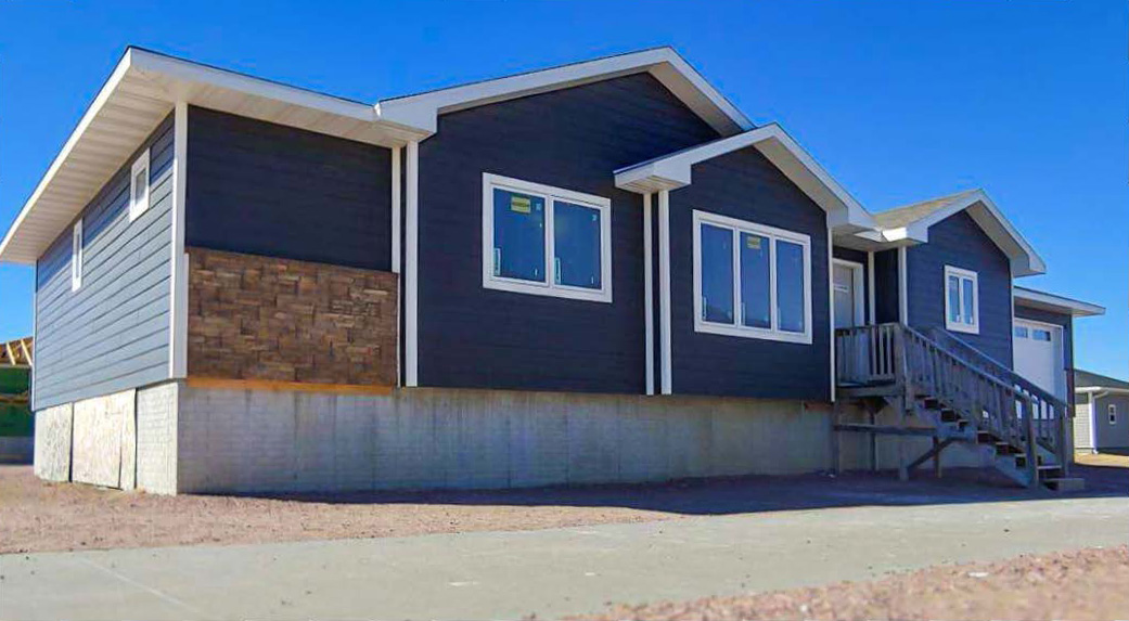 Side exterior view of a single-story house with dark blue siding, white trim, multiple gabled rooflines, raised concrete foundation, and exterior stairs leading to an entry deck.