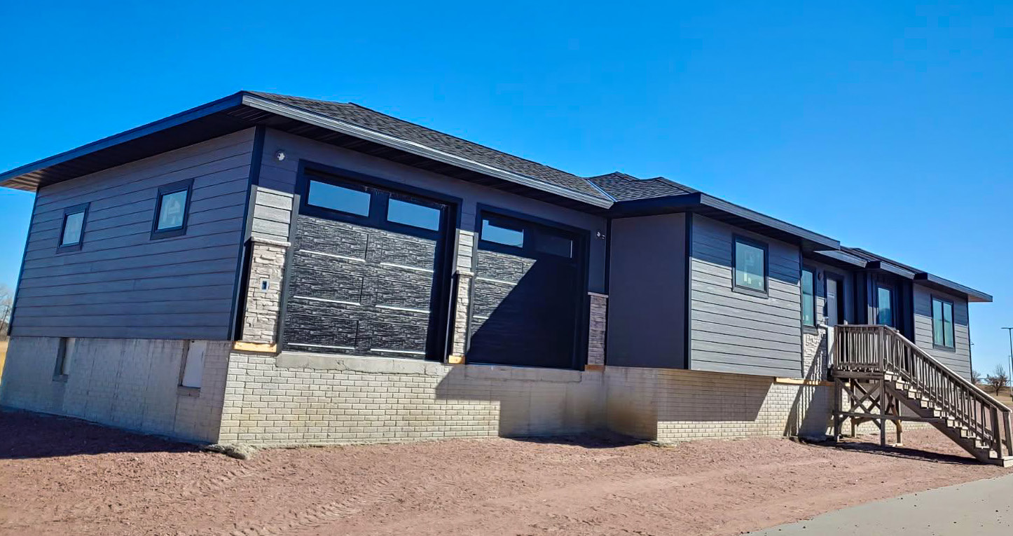 Front exterior of a single-story modern house with gray siding, stone accents, attached garage, and a raised foundation under a clear blue sky.