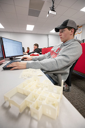 Person seated at a desktop computer in a classroom, using design software, with a white 3D‑printed building model in the foreground.