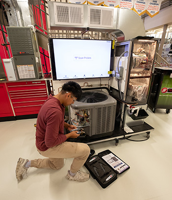 HVAC apprentice kneels beside an open air conditioning condenser in a training lab, using diagnostic tools while instructional equipment and ductwork are visible behind them.